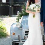 Femme vêtue d'une robe de mariée cymbeline avec un bouquet au bras de son mari devant une voiture ancienne